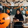 Eine Frau sitzt in einem herbstlich geschmückten Klassenraum und liest eine Halloween-Geschichte vor. Bunte Kürbisse, gebastelte Hexen und herbstliche Deko machen deutlich: So schön, warmherzig und ganz ohne Grusel kann eine Lesung zum Herbstfest sein – passend zu „Die Kürbishexe“.