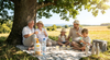 Mehrgenerationen-Familie sitzt an einem hei&szlig;en Sommertag entspannt im Schatten eines Baumes mit Erfrischungsgetr&auml;nken und Sonnencreme &ndash; wirksamer Hitzeschutz f&uuml;r die Gesundheit im Alltag